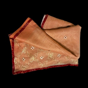 May include: Peach-colored scarf with gold floral embroidery and red trim. The scarf has a striped pattern and small red and white accents. The edges are finished with a patterned red border.