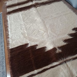 May include: A rectangular, shaggy rug with a cream-colored background and dark brown geometric patterns. The rug has a fringe along the edges and is made of a soft, textured material. The design features stripes and irregular shapes.
