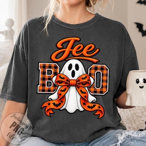 Może przedstawiać: Ciemnoszary t-shirt z grafiką białego ducha noszącego pomarańczowo-czarną kokardę w nietoperze. Napis "Jee BO" jest nadrukowany pomarańczowo-czarnymi literami w kratę, z dodatkowym pomarańczowym napisem wykonanym ozdobną czcionką.