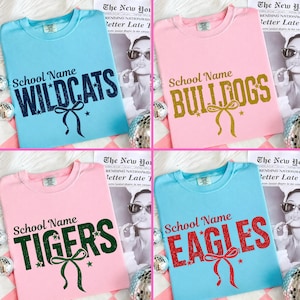 May include: A collection of four short-sleeved crewneck t-shirts in light blue and light pink. Each shirt features the text School Name followed by a team name in a distressed font, including WILDCATS, BULLDOGS, TIGERS, and EAGLES with a bow.