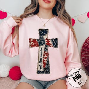 Op de afbeelding: Een lichtroze sweatshirt met een kruisontwerp gemaakt van rode, witte en blauwe patches, die lijken op de Amerikaanse vlag. De woorden "god bless america" staan verticaal op het kruis gedrukt. Een casual top.