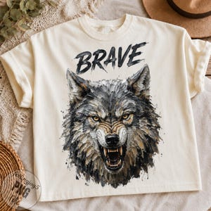Puede incluir: Una camiseta de manga corta color crema con un gráfico de una cabeza de lobo gruñendo con pelaje texturizado estilo pincelada en tonos gris, negro y tostado. La palabra BRAVE está impresa en una fuente negra, negrita y desgastada.