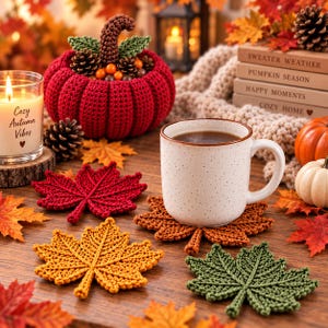 May include: A set of three crocheted maple leaf coasters in red, yellow, and green, displayed alongside a white ceramic mug, a red crocheted pumpkin, a candle labeled Cozy Autumn Vibes, and a stack of books titled Sweater Weather and Pumpkin Season.