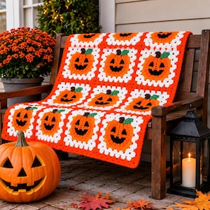Può includere: Una coperta all'uncinetto con un motivo ripetuto di quadrati arancioni a forma di zucca con steli verdi e tratti del viso neri, bordati con filato bianco e arancione. La coperta è drappeggiata su una panca di legno.