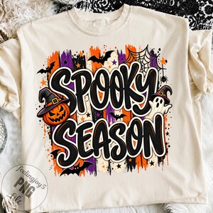 Könnte beinhalten: Ein cremefarbenes T-Shirt mit einem Grafikdruck, der den Text "SPOOKY SEASON" in großen schwarzen Buchstaben zeigt. Das Design umfasst einen Kürbis mit Hexenhut, ein Gespenst, Fledermäuse, ein Spinnennetz sowie orange und violette Pinselstriche.
