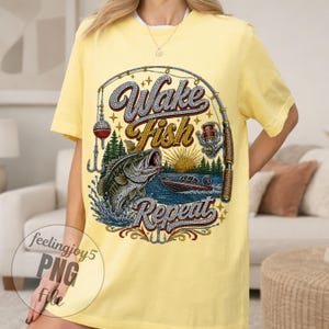 May include: Yellow t-shirt with a fishing-themed graphic. The design features a fish jumping out of the water, a fishing rod, and the words "Wake Fish Repeat" in a decorative font. A boat is also visible.