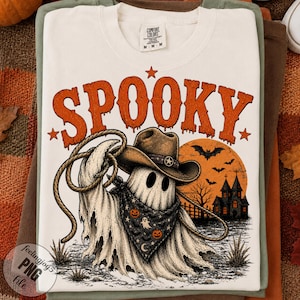 May include: A cream-colored t-shirt featuring the orange text SPOOKY above a graphic of a ghost wearing a cowboy hat and a patterned bandana. The design includes a lasso, a large orange moon, a haunted house, and bats, all printed on the front.