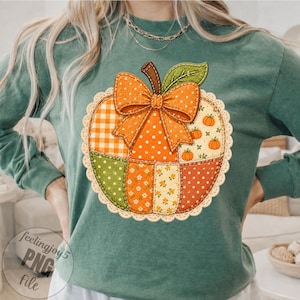 Puede incluir: Una camiseta de manga larga color verde salvia con un gráfico central de una calabaza de patchwork. La calabaza está compuesta por varios patrones de tela en naranja, verde y crema, decorada con un lazo naranja de lunares y un borde de encaje.