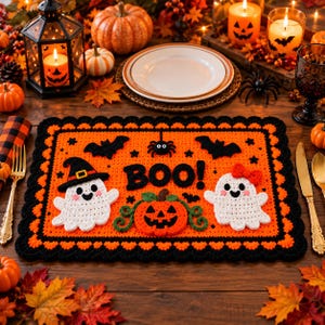 May include: A rectangular crochet placemat featuring an orange background with a black scalloped border. The design includes two white ghosts, one wearing a black hat and one with a red bow, a central orange jack-o'-lantern, and the black text BOO!
