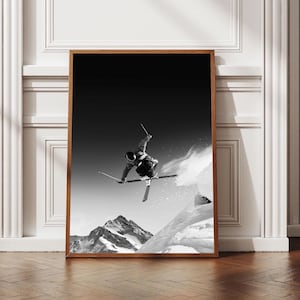 May include: A black and white framed wall art print featuring a skier mid-air during a jump. The skier is captured against a dark sky, positioned above snow-covered mountain peaks. The frame is made of wood with a natural brown finish.
