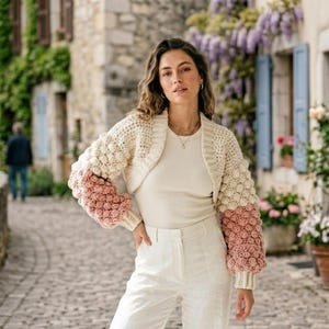 Könnte beinhalten: Cremefarbenes Häkel-Bolero mit Blasenärmeln in Creme und Staubrosa. Der kurze Pullover hat einen runden Ausschnitt. Die Ärmel sind lang und enden am Handgelenk. Getragen über einem weißen Oberteil.
