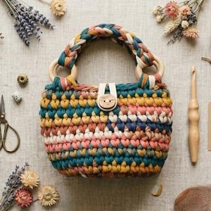 May include: A crocheted handbag featuring a multi-colored striped pattern in teal, mustard yellow, and dusty pink. The bag has a braided handle, two wooden ring attachments, and a central wooden button closure with a leather tab. A tag reads HANDMADE WITH LOVE.