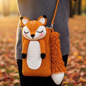 May include: A crocheted orange fox-shaped crossbody bag with a long, fluffy tail. The bag features a white belly, black paws, and a fox face with closed eyes and pink cheeks. The strap is orange.