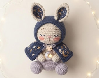 Könnte beinhalten: Ein gehäkeltes Stoffhasenspielzeug aus cremefarbenem Garn, das einen dunkelblauen Kapuzenumhang mit goldenen Mond- und Sternenmustern trägt. Der Hase hat geschlossene Augen, rosa Wangen und hält eine kleine klare Kugel mit einem Stern sowie ein Schild mit Boo!
