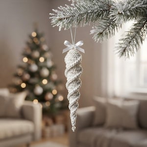 May include: A white crocheted icicle ornament with a spiral texture, featuring a small white satin ribbon bow at the top and a silver hanging cord. The ornament tapers to a point at the bottom, finished with a small clear bead detail.