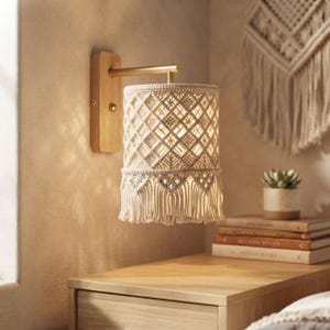 May include: A wall-mounted lamp featuring a rectangular light-colored wooden base and a cylindrical lampshade crafted from off-white macrame cord. The shade displays a geometric diamond pattern and terminates in a fringe of hanging cords.