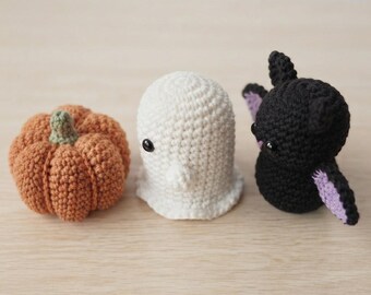 Könnte beinhalten: Drei kleine gehäkelte Figuren, bestehend aus einem orangefarbenen Kürbis mit grünem Stiel, einem weißen Geist mit schwarzen Perlenaugen und einer schwarzen Fledermaus mit violetten Flügelinnenseiten und schwarzen Perlenaugen.