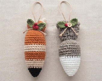 Könnte beinhalten: Zwei gehäkelte Ornamente in Form von Tierschwänzen mit Aufhängeschlaufen. Der linke Schwanz ist orange, cremefarben und schwarz mit grünen Blättern und roten Beeren. Der rechte Schwanz ist grau und weiß mit grünen Blättern und einem Holzknopf.