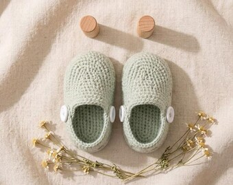 Könnte beinhalten: Ein Paar hellsalbeigrüne gehäkelte Babyschuhe mit abgerundeter Spitze, tiefer Öffnung und einem weißen Knopf an jeder Seite. Zwei kleine Holzzylinder sind über den Schuhen platziert, und Zweige mit getrockneten weißen Blumen liegen unterhalb.