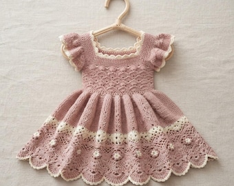 Könnte beinhalten: Ein altrosa gehäkeltes Kleid mit quadratischem Ausschnitt, gerüschten Flügelärmeln und einem ausgestellten Rock. Das Oberteil hat ein strukturiertes Muster, während der Saum mit einer breiten, cremefarbenen Spitzenborte mit Blumenmotiven verziert ist.