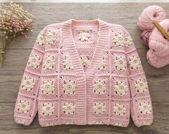 Könnte beinhalten: Eine langärmelige Strickjacke aus gehäkelten Quadraten in Hellrosa und Creme. Jedes Quadrat zeigt ein cremefarbenes Blumenmotiv in der Mitte, umgeben von rosa Garn. Das Kleidungsstück hat einen V-Ausschnitt, eine knopflose Vorderseite und Rippbündchen.