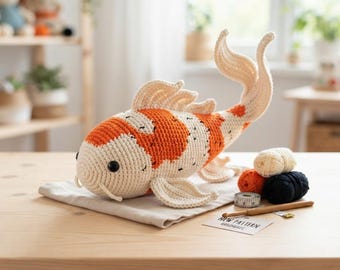 Könnte beinhalten: Ein gehäkeltes Koi-Fisch-Spielzeug mit cremefarbenem Körper, orangefarbenen Flecken und schwarzen Sprenkeln. Der Fisch hat schwarze Knopfaugen, ausgeprägte Flossen und einen langen Schwanz. Daneben liegen kleine Wollknäuel, ein Maßband und eine Häkelnadel.