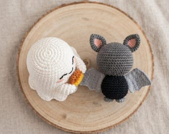 Könnte beinhalten: Eine weiße gehäkelte Geisterfigur, die einen kleinen orangefarbenen Kürbis hält, und eine grau-schwarze gehäkelte Fledermausfigur mit hellrosa Innenohren und hellgrauen Flügeln, beide mit deutlich sichtbaren Maschen gefertigt.