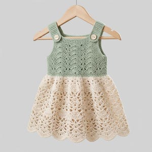 May include: A sleeveless dress featuring a sage green crocheted bodice with a scalloped pattern and two light-colored wooden buttons at the shoulders. The skirt is made of cream-colored crocheted fabric with an intricate, repeating fan-like design.