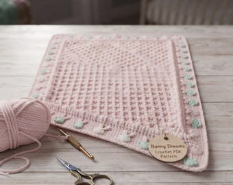 Könnte beinhalten: Eine rechteckige, hellrosa gehäkelte Decke mit Waffelmuster und einer Bordüre aus kleinen, mintgrünen Hasenmotiven. Ein Knäuel passendes rosa Garn, eine goldene Häkelnadel, eine Schere und ein rundes Holzschild mit der Aufschrift Bunny Dreams Crochet PDF Pattern liegen daneben.