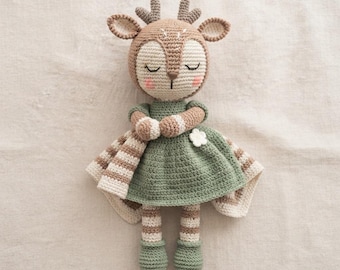Könnte beinhalten: Eine gehäkelte Rehkitz-Puppe mit beigem Kopf, geschlossenen Augen, rosa Wangen und kleinen Geweihen. Die Puppe trägt ein salbeigrünes Kleid mit einer weißen Blumenapplikation sowie gestreifte Arme und Beine in Beige und Weiß mit salbeigrünen Füßen.