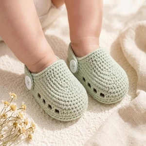 May include: A pair of light sage green crochet baby shoes featuring a closed-toe design with small side cutouts and a single white button closure on the outer side of each shoe. The footwear is crafted with a textured yarn stitch pattern.