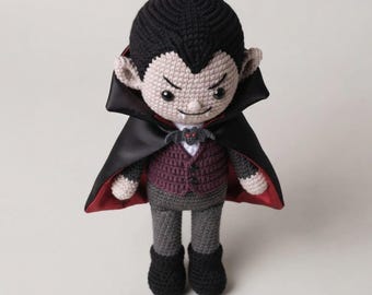 Könnte beinhalten: Eine gehäkelte Vampirpuppe mit schwarzem Haar, blasser Haut und zwei kleinen weißen Fangzähnen. Die Puppe trägt eine dunkelviolette Weste, eine graue Hose, schwarze Schuhe und einen schwarzen Satinumhang mit rotem Futter und einer kleinen Fledermausbrosche.