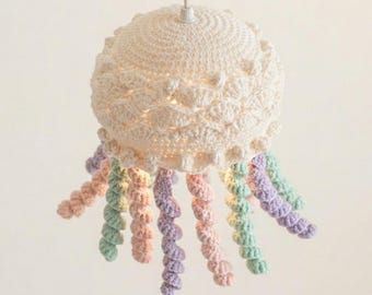 Könnte beinhalten: Eine cremefarbene, gehäkelte Pendelleuchte in Form einer Qualle. Die Kuppel weist ein strukturiertes Muschelmuster auf, während zahlreiche spiralförmige Tentakel in Pastelltönen wie Rosa, Violett und Mintgrün vom unteren Rand herabhängen.