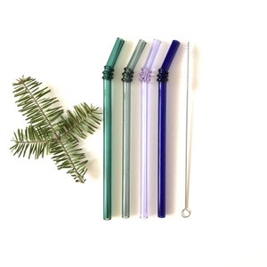 May include: Four reusable glass straws in shades of green, grey, and purple, plus a blue one, are displayed on a white background. A stainless steel cleaning brush and a sprig of greenery are also present. The straws have a decorative spiral design.