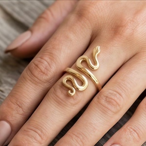 May include: A gold-toned ring shaped like a coiled snake that wraps around the finger. The metal has a smooth, polished finish, and the design features the head of the snake at one end and a curved tail at the other, creating an open-ended band.
