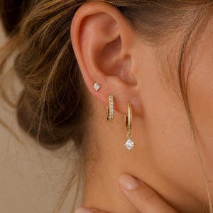 May include: A close-up of a person's ear adorned with three gold earrings. The set includes a small square stud, a small hoop with embedded crystals, and a larger hoop with a dangling square crystal. The earrings are set against the person's skin.