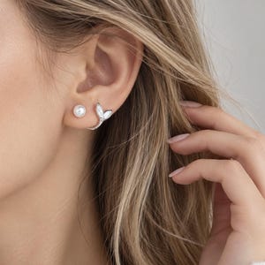 May include: A silver-toned earring set consisting of a small, round white pearl stud and a separate silver-toned earring shaped like a curved mermaid tail or leaf, both worn on the earlobe. The jewellery features a polished metallic finish.