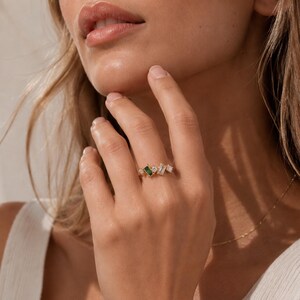 May include: A gold ring featuring an emerald-cut green gemstone, flanked by several white rectangular and square-cut stones. The ring is set on a hand with a neutral background.