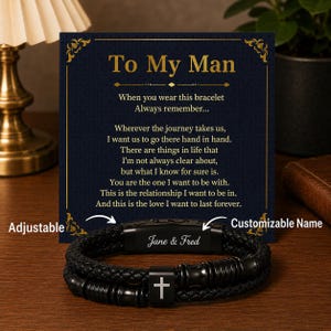 Könnte beinhalten: Schwarzes Lederarmband mit Kreuzanhänger und den eingravierten Namen "Jane & Fred". Das Armband ist verstellbar und hat ein geflochtenes Design. Eine Karte mit der Aufschrift "To My Man".