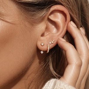 May include: An assortment of gold earrings, including a small hoop, a stud with a single diamond, a diamond-shaped stud, and a cluster of three small diamonds. The earrings are displayed on an ear.