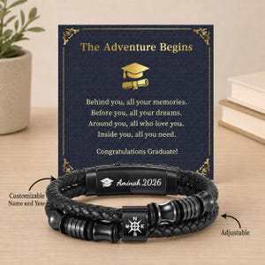 Pode incluir: Pulseira de couro trançado preta com design de bússola e o texto "Aminah 2026". A pulseira é ajustável e vem com um cartão que diz "A Aventura Começa" e "Parabéns Graduado!"