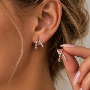 May include: A silver-toned earring featuring a smooth, curved metal arch paired with a smaller, straight bar encrusted with five small, clear, sparkling stones. The earring is designed to wrap around the earlobe, creating a layered, dual-piece appearance.