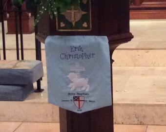 Könnte beinhalten: Ein hölzerner Lesepult mit einem blauen Banner, auf dem "Erik Christopher" steht und ein weißes Lamm mit den Worten "Heilige Taufe" darunter. Das Banner enthält auch ein Wappen mit einem roten Kreuz und den Worten "Kirche St. Marien".