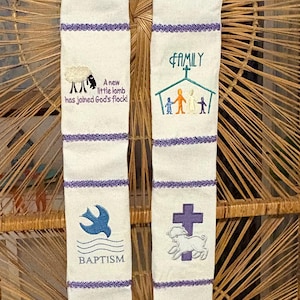 May include: Two white fabric panels with purple trim and embroidered religious imagery. The left panel features a dove, cross, and lamb with the text "A new little lamb has joined God's flock!" and "Baptism". The right panel features a seashell, a cross, a house with a family inside, and the text "Family" and "He's got the whole world in his hands".