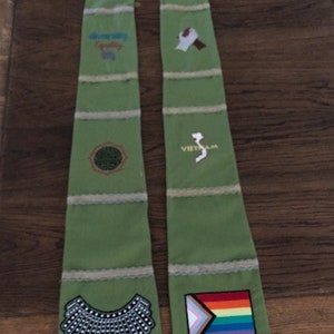 K&ouml;nnte beinhalten: Ein gr&uuml;nes Stola mit gestickten Aufn&auml;hern. Zu den Aufn&auml;hern geh&ouml;ren ein schwarz-wei&szlig;er Kragen, eine Regenbogenflagge und der Text "Vietnam". Die Stola hat horizontale beige Streifen mit Spitzensaum.