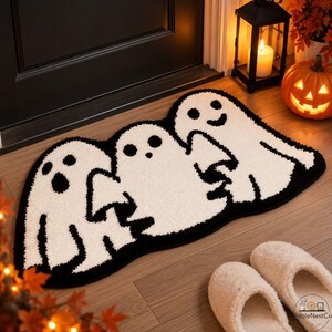 Op de afbeelding: Een wit, spookvormig vloerkleed met zwarte omlijningen en gelaatstrekken, waarop drie afzonderlijke spookfiguren met elkaar verbonden zijn. Het kleed heeft een zacht, getextureerd oppervlak en een zwarte rand die de contouren volgt.