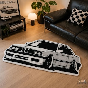 May include: A black and white car-shaped rug. The rug features a detailed outline of a classic car, with black tires, windows, and accents against a gray body. The rug is placed on a wooden floor.