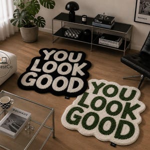 May include: Two decorative rugs with the phrase "YOU LOOK GOOD" in bold letters. One rug is black with white lettering, and the other is off-white with green lettering. The rugs are on a wooden floor.