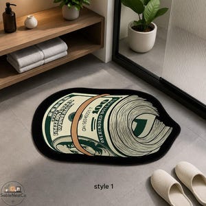 May include: An oval-shaped floor mat featuring a graphic print of a rolled-up one hundred dollar bill. The design includes the text "THE UNITED STATES OF AMERICA" and "ONE HUNDRED DOLLARS" with a black border surrounding the entire mat.