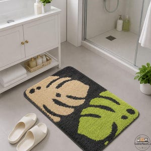 May include: A rectangular bath mat featuring a dark grey background and large leaf designs in tan and green. The mat is on a light grey tiled floor near a white vanity and shower.
